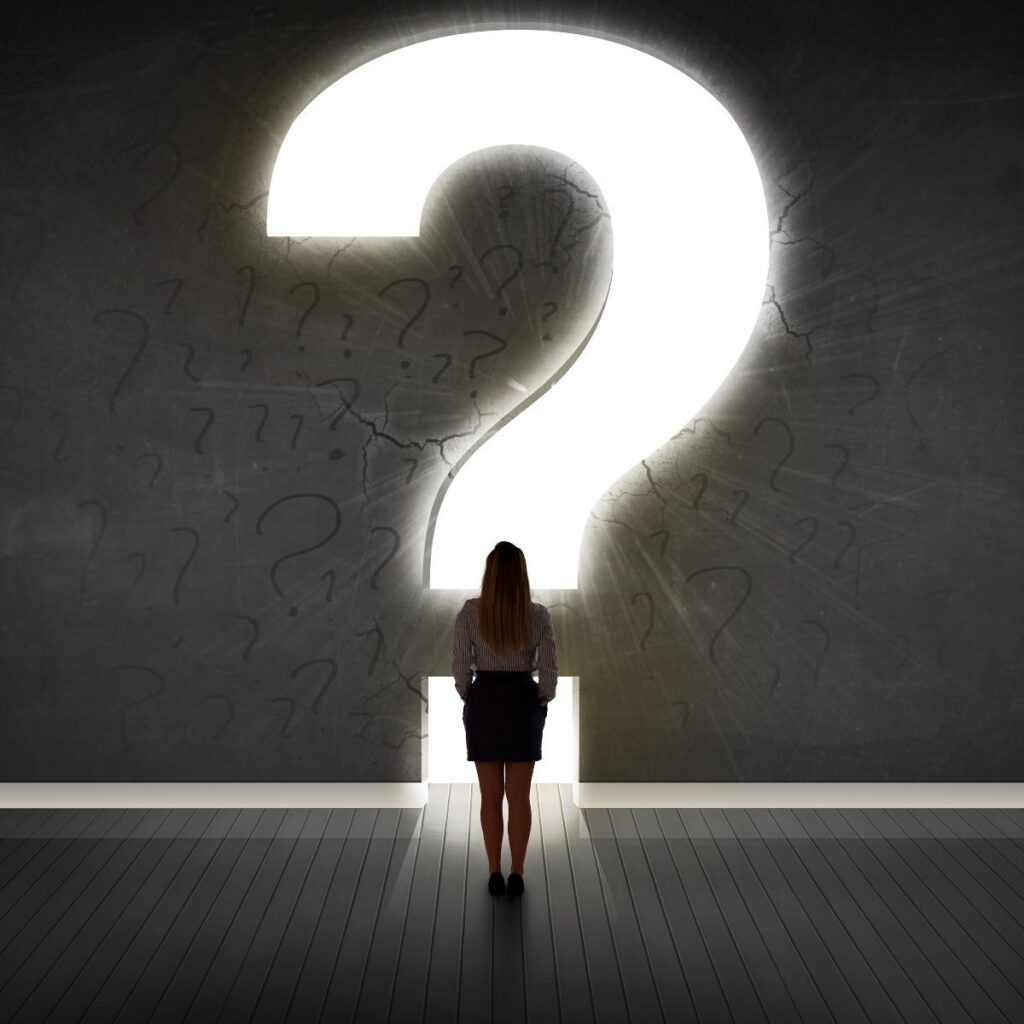 A person standing in front of a large glowing question mark on a wall filled with smaller question marks, suggesting uncertainty and exploration.