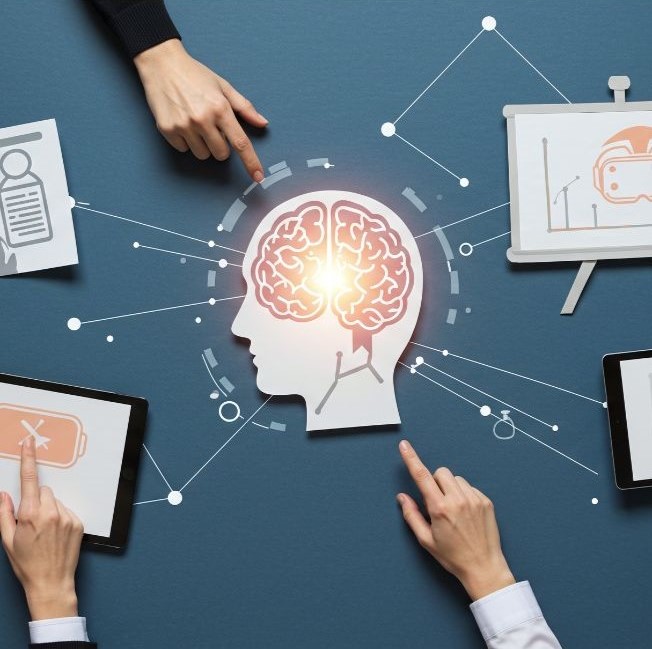 Multiple hands pointing toward a central brain icon on a table, with surrounding devices displaying charts, documents, and graphs, showing collaboration and shared decision-making around data.