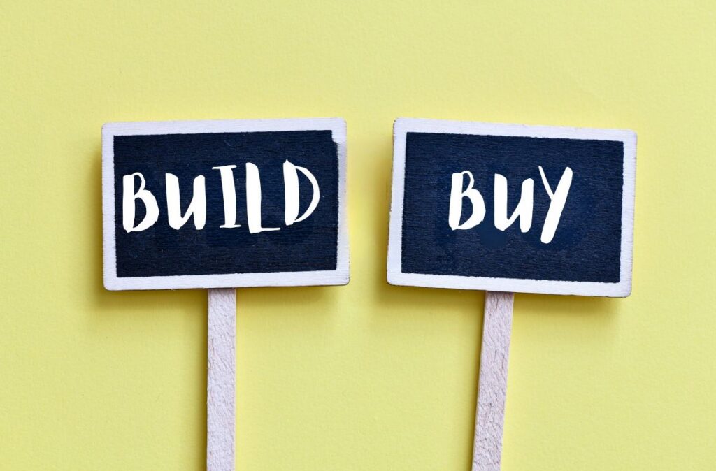 Two small signboards labeled “BUILD” and “BUY” placed side by side on a yellow background, representing a decision between creating or purchasing a solution.