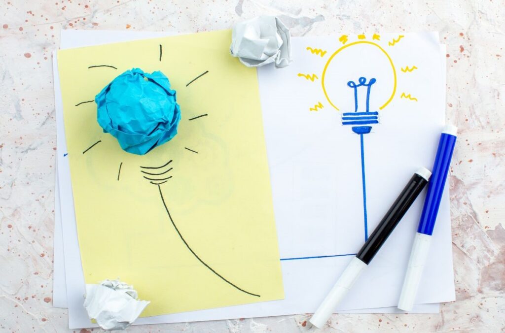 Two sketches of light bulbs on paper, one made from a crumpled paper ball and the other drawn clearly with markers, representing the progression from a rough idea to a refined concept.
