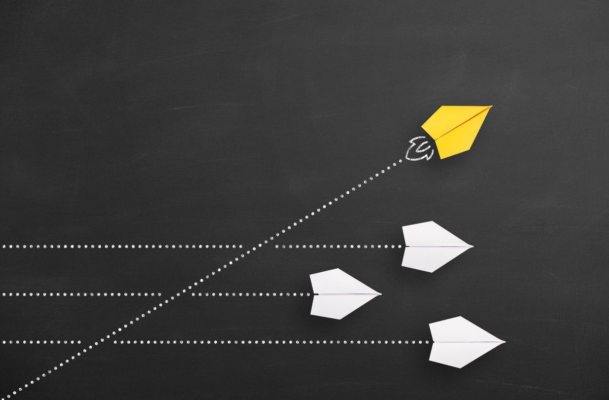 Yellow paper airplane diverging upward from a group of white paper airplanes following straight paths, illustrating differentiation, innovation, or breaking away from the norm.